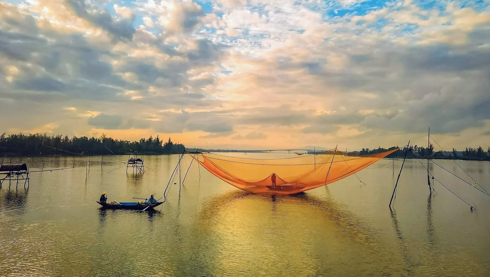 A giant fishing net glows orange under a golden sunset sky on a river near Hoi An.