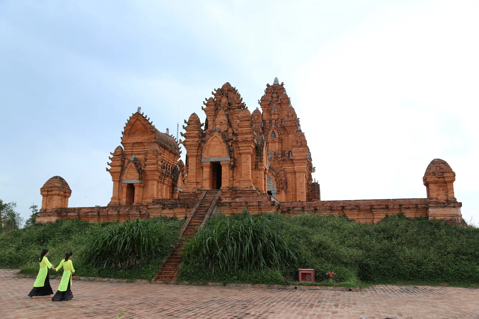 Two women in yellow ao dai walk toward an ancient red brick Cham tower temple in Vietnam.