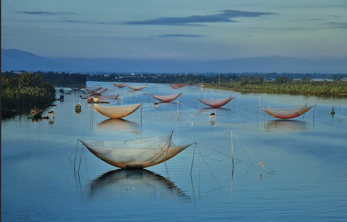Traditional square lift-nets dot a calm river at dusk in the Hoi An countryside.