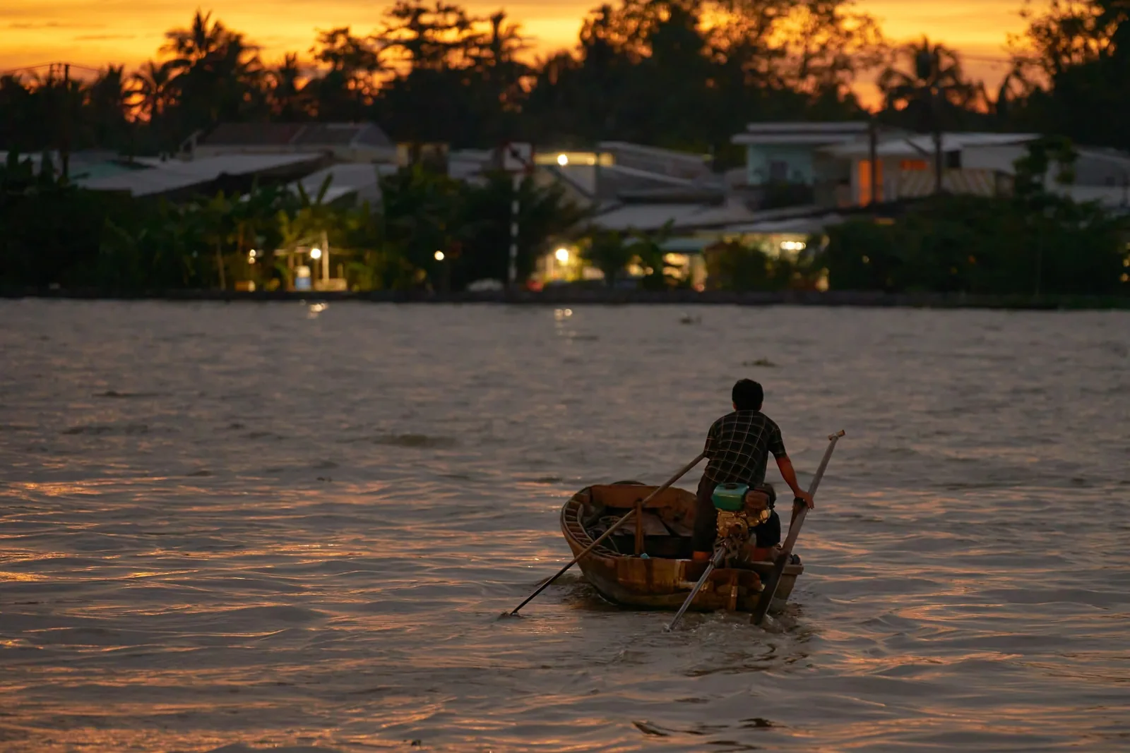 A man rows a small wooden boat across the Mekong Delta river at dusk with village lights glowing on the far bank.