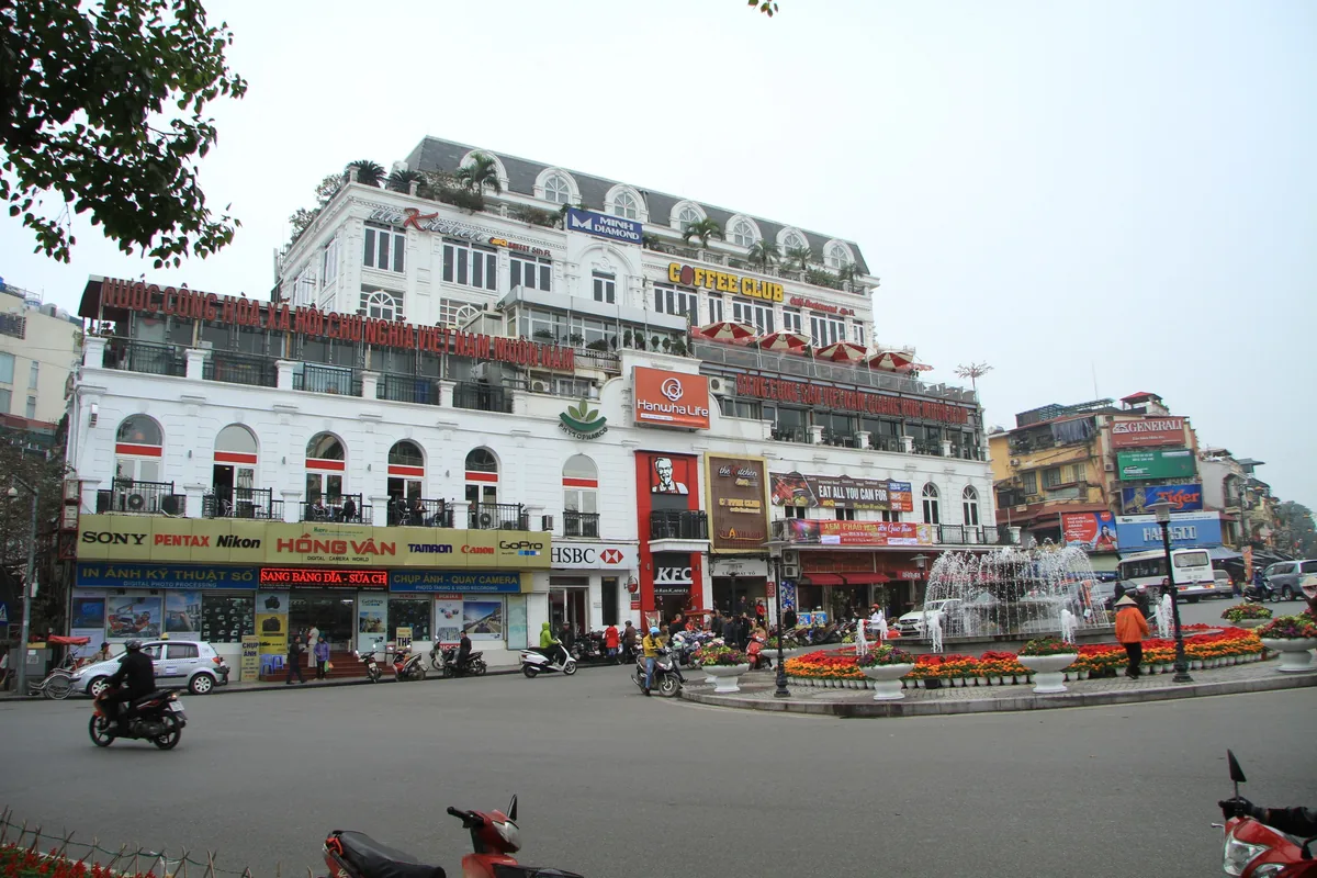 A busy Hanoi street corner with a fountain, flower beds, motorbikes and shophouses covered in colorful signage.